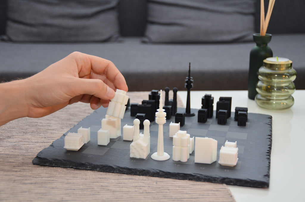 Person spielt München Design-Schach: Hand bewegt weiße Skyline-Figur auf dem lasergravierten Naturschiefer-Brett im Wohnzimmer.