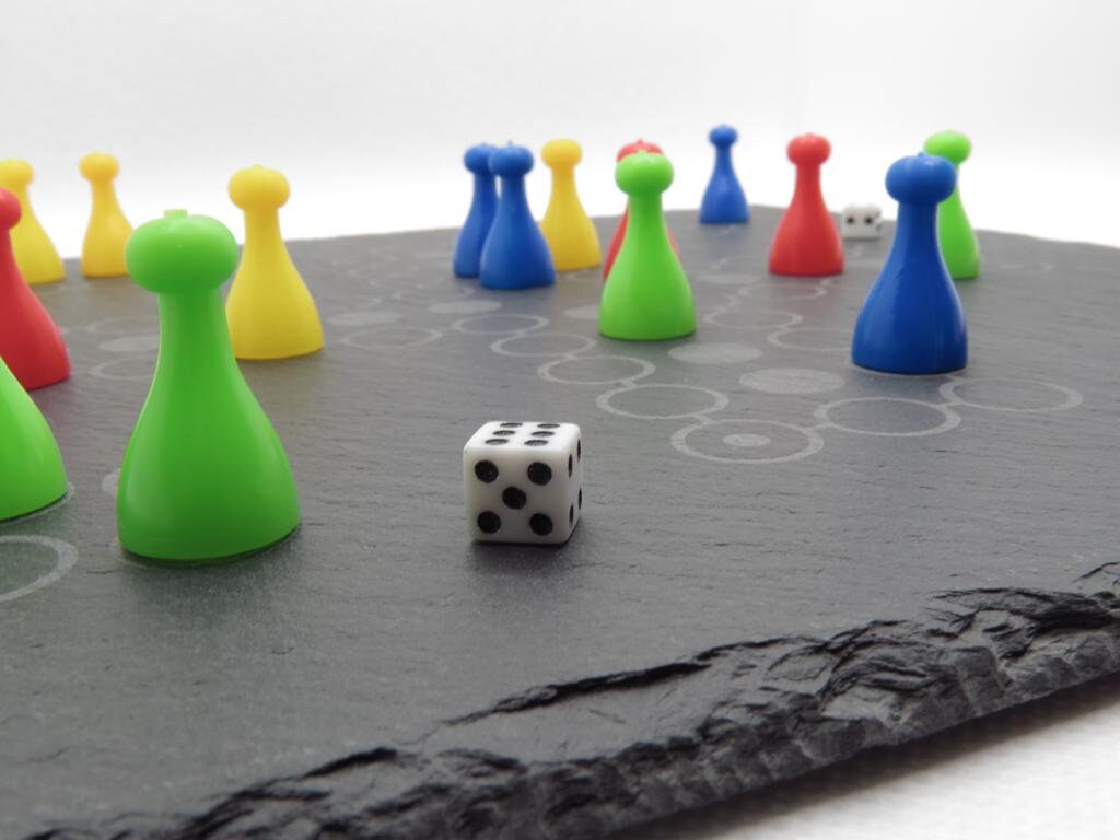 Ludo Spielsituation auf Schiefer: Bunte Spielfiguren und Würfel auf dem edlen grauen Steinbrett.