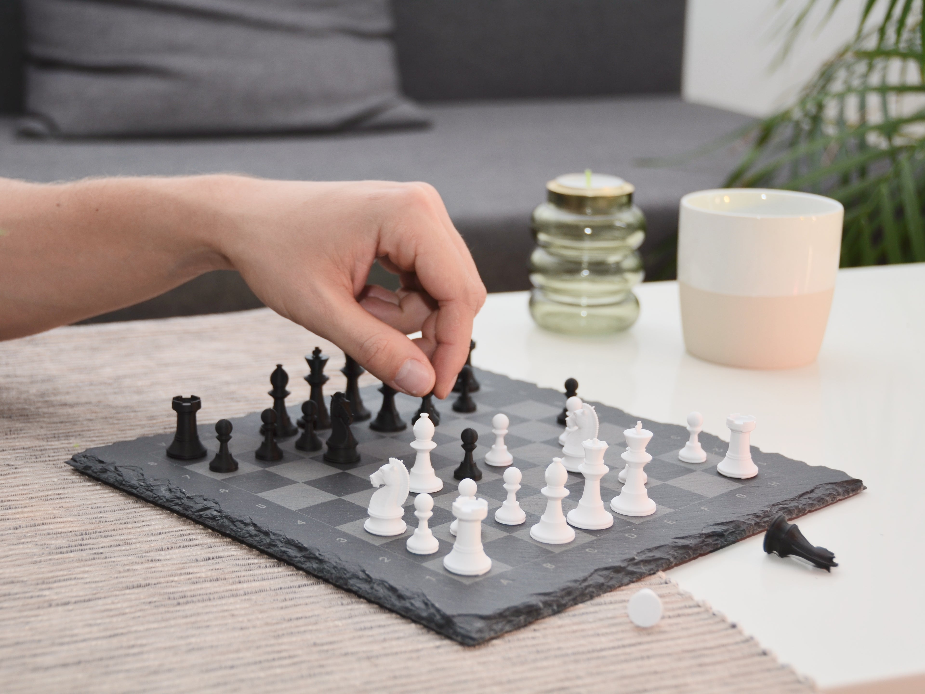 Person spielt Schach am Couchtisch: Hand bewegt weiße Bauern-Figur auf dem gravierten Schiefer-Schachbrett.